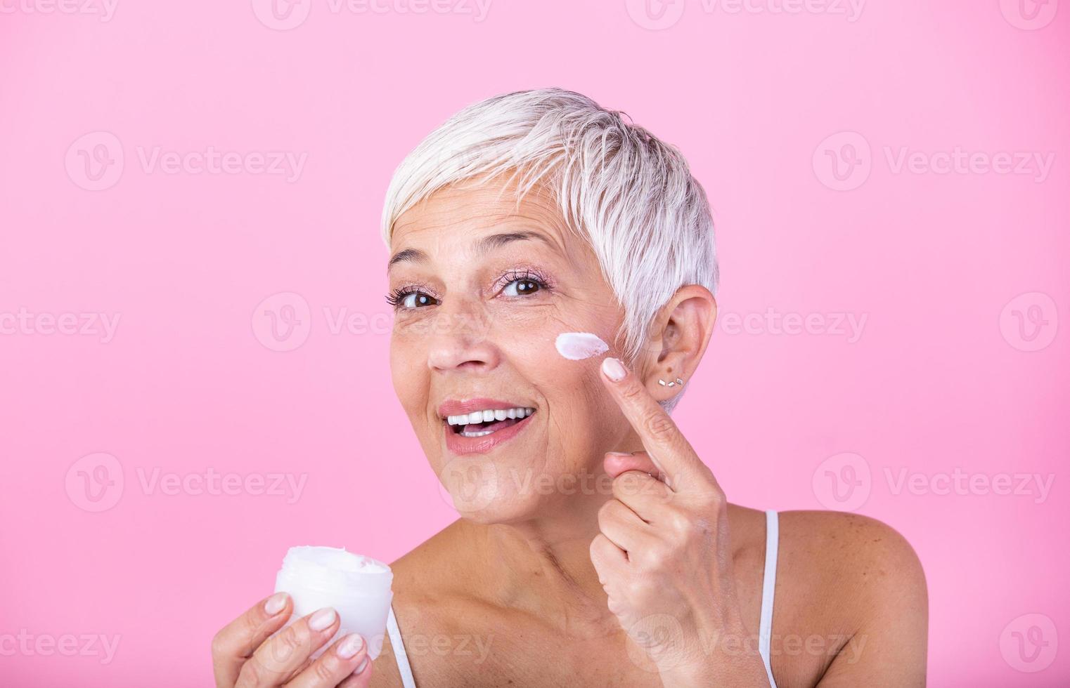 Beautiful mature woman holding jar of moisturizer and looking at camera. Happy senior woman holding a bottle of anti-aging lotion isolated over pink background. Anti aging and beauty treatment. photo