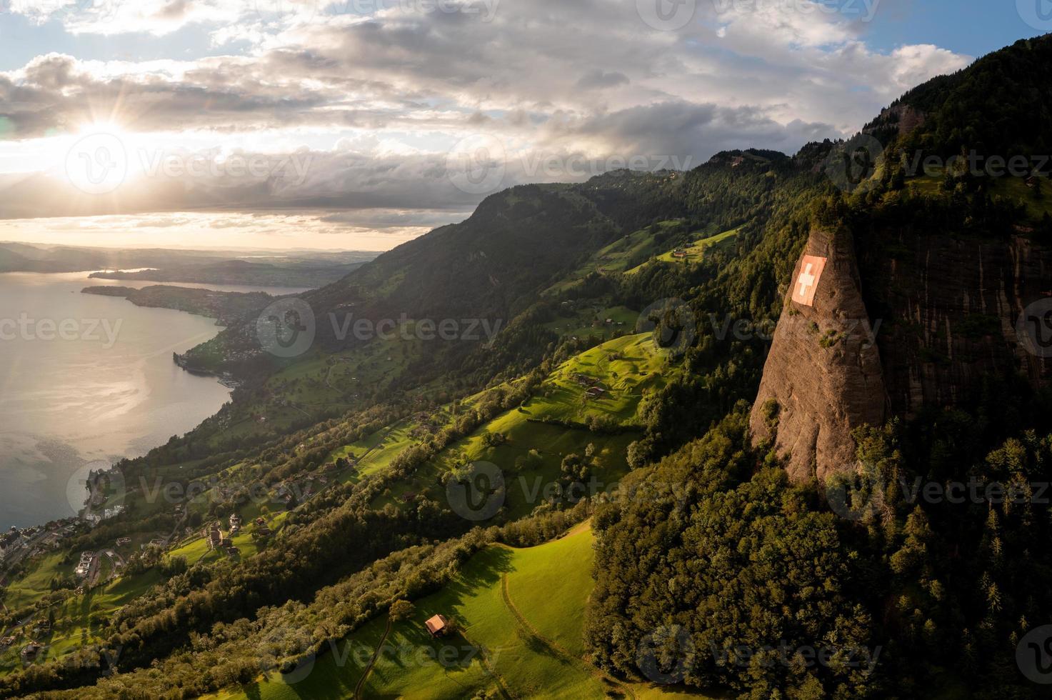 a huge swiss flag is fixed on a cliff, below it is a village on a lake photo