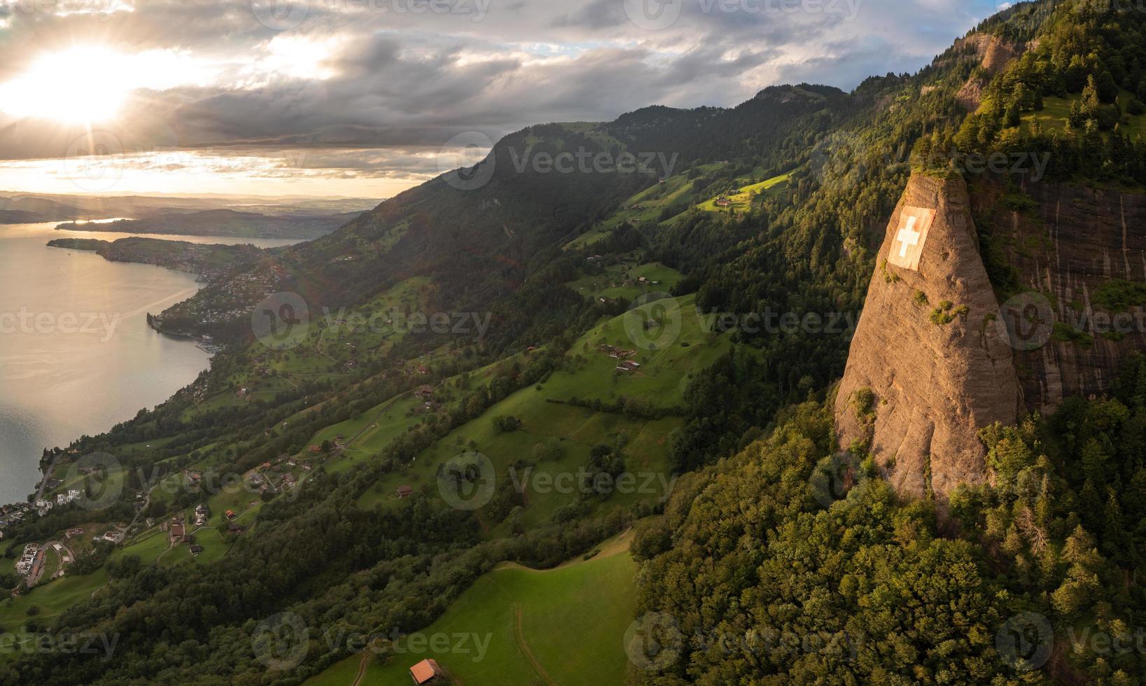 a huge swiss flag is fixed on a cliff, below it is a village on a lake photo