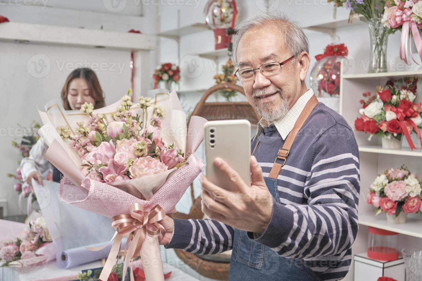 Happy Asian senior male florist owner selfie pictures and show floral bouquet arrangement with smartphone in valentine at bright flower shop, beautiful blossoms store, small business SME entrepreneur. photo