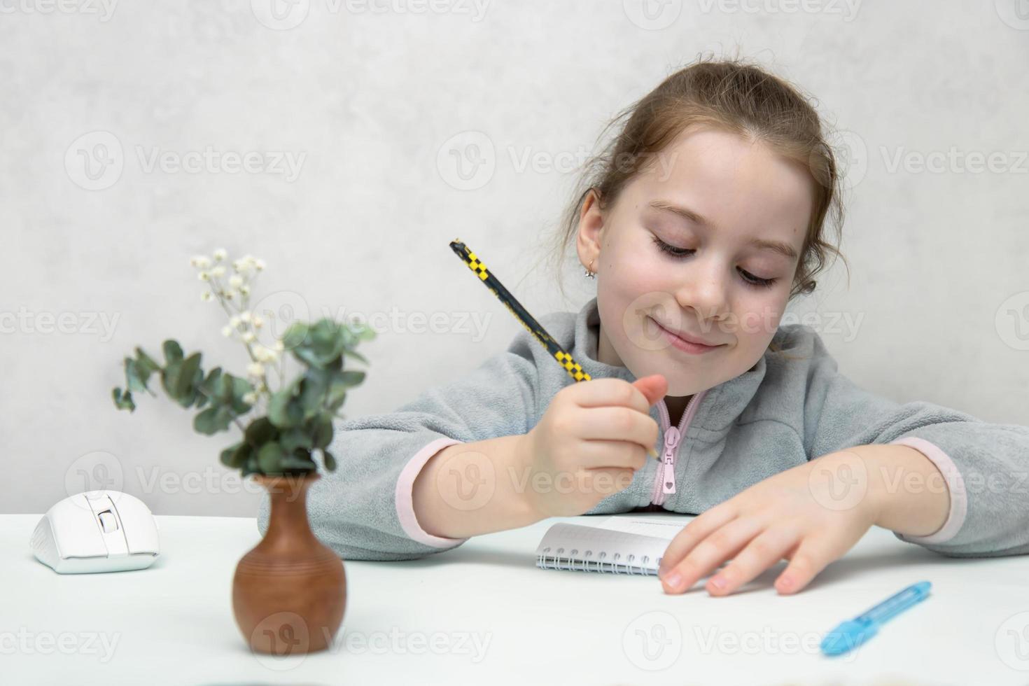 cute little girl sitting at the table smiling writing with a pencil in a notebook, learning, back to school photo