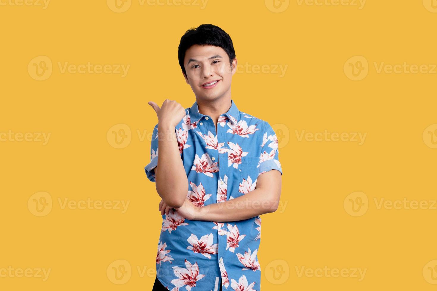 Young Asian man in blue floral shirt smiling and pointing hand sideways on yellow background photo