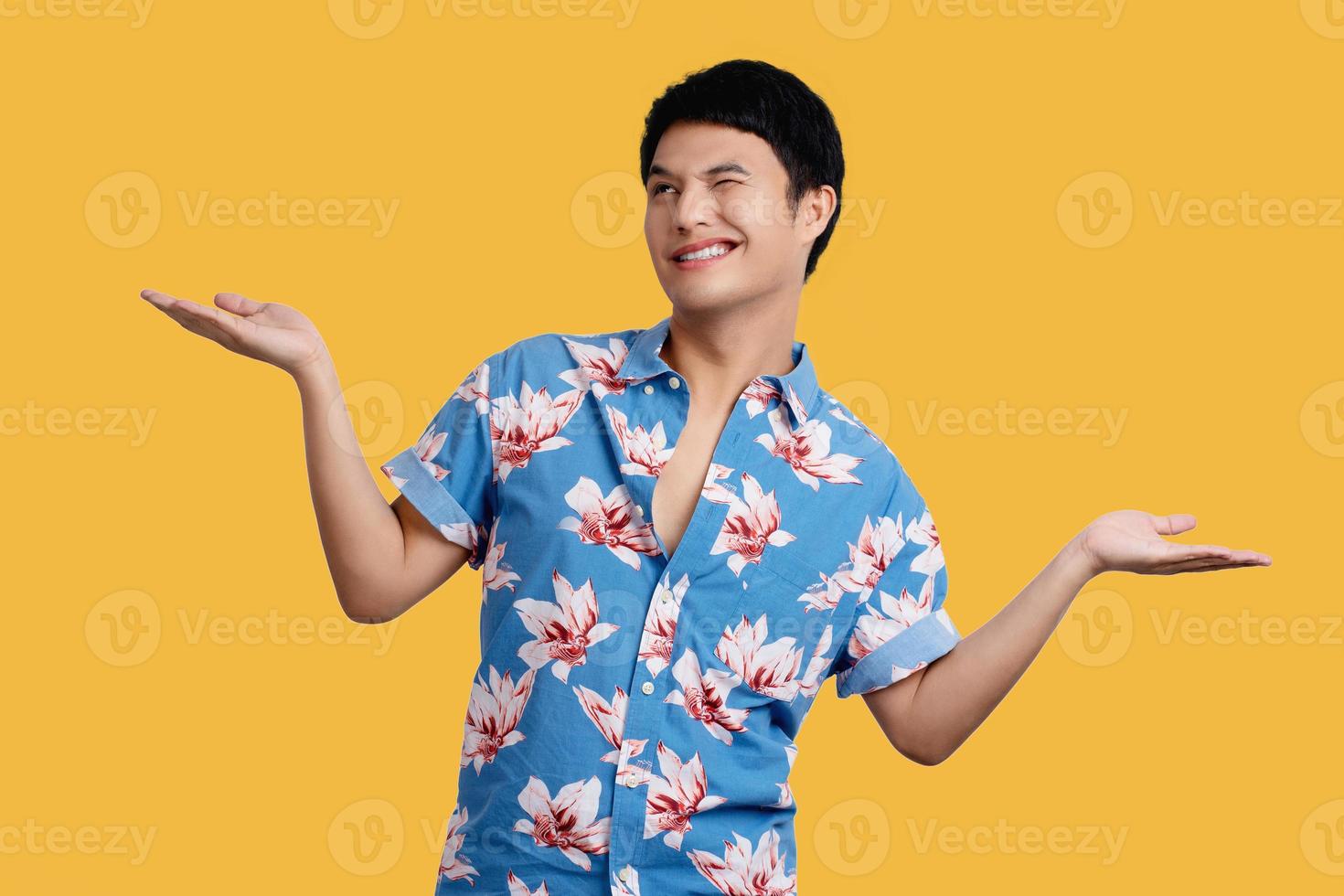 Smiling young handsome Asian man  presenting with open palms gesture on yellow background in studio. He in blue floral shirt for Songkran festival in Thailand. photo