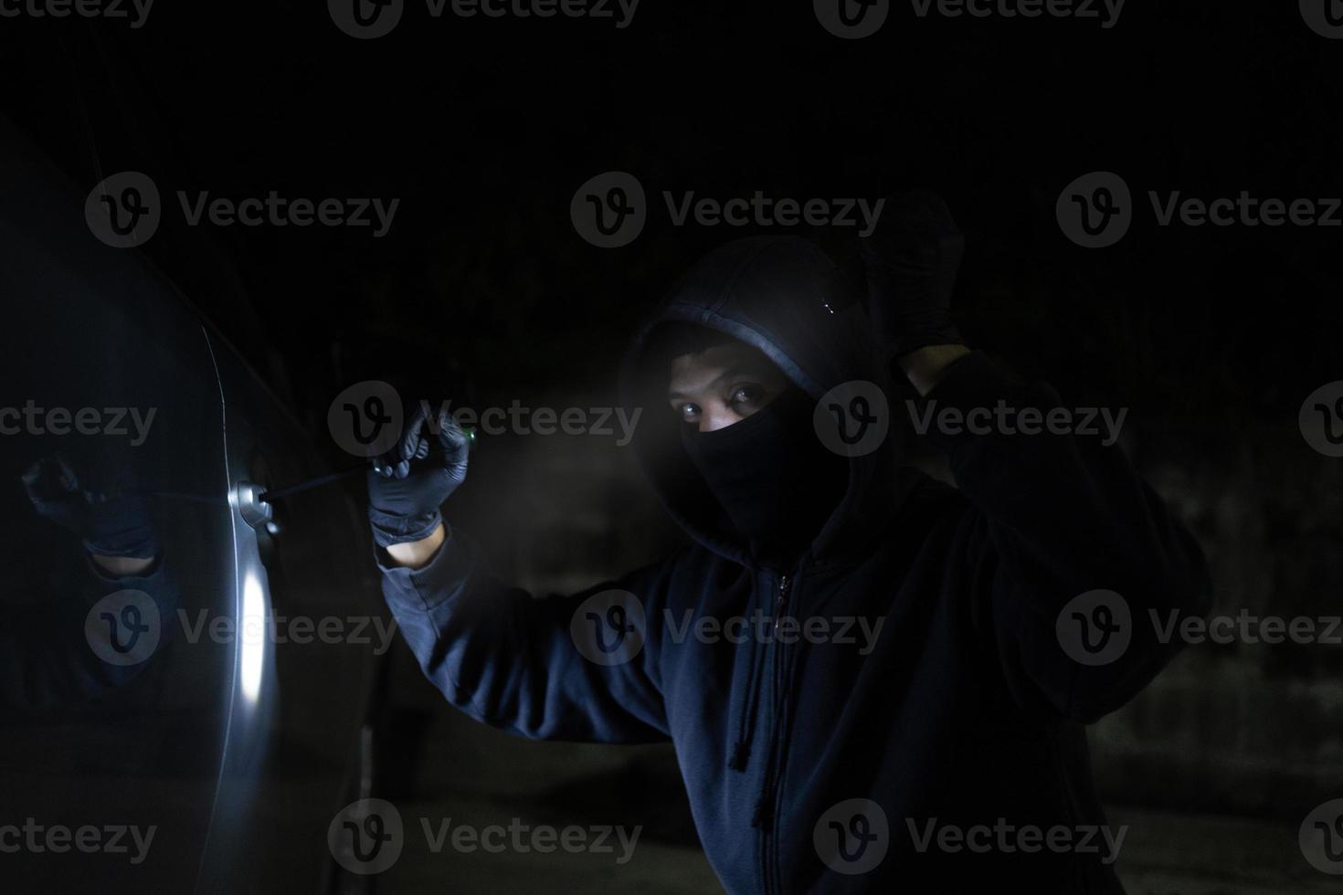 Young thief in black is stealing a car at night. He carries a flashlight and a screwdriver for car theft. photo