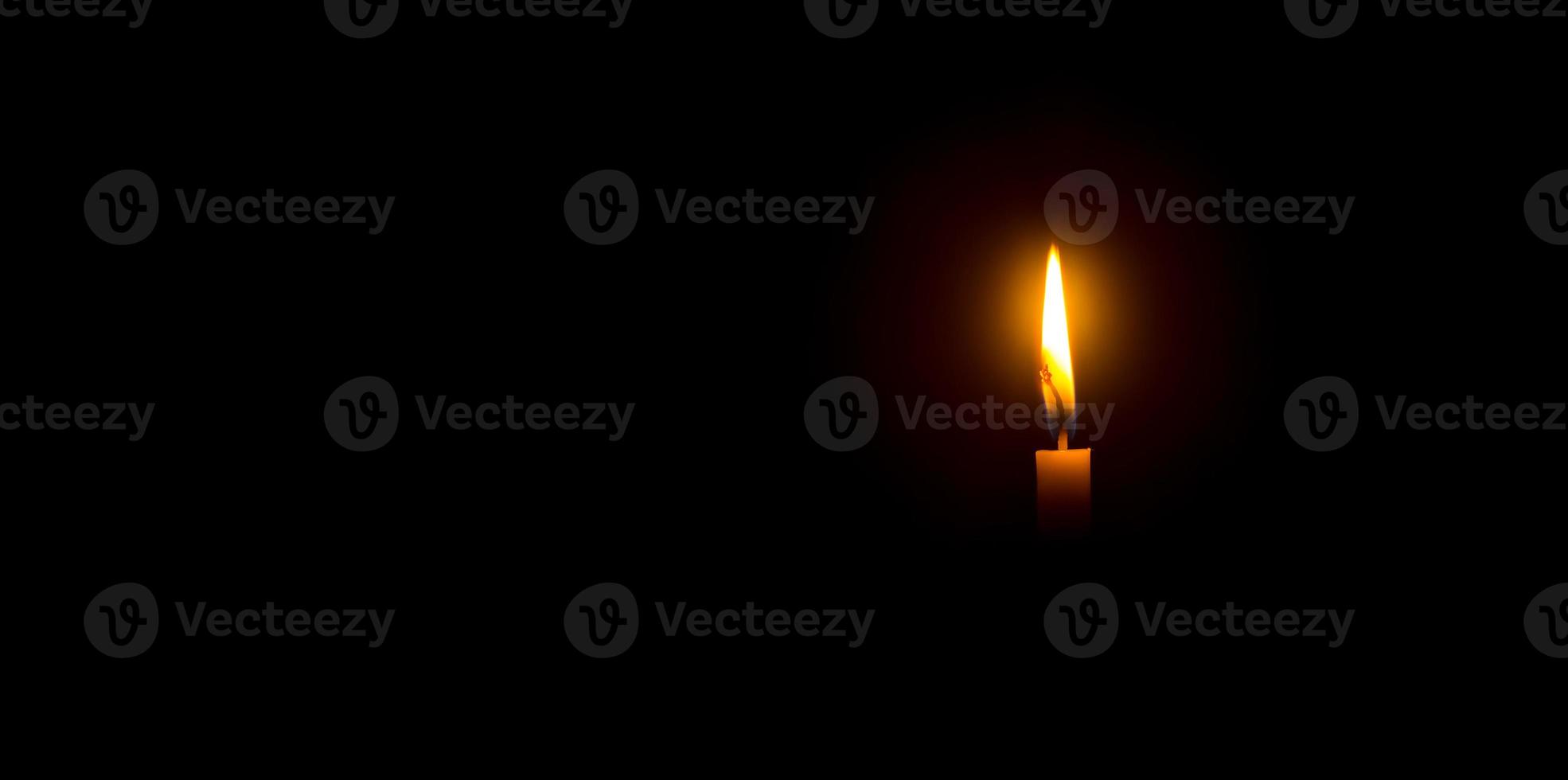 A single burning candle flame or light glowing on an orange candle on black or dark background on table in church for Christmas, funeral or memorial service with copy space photo