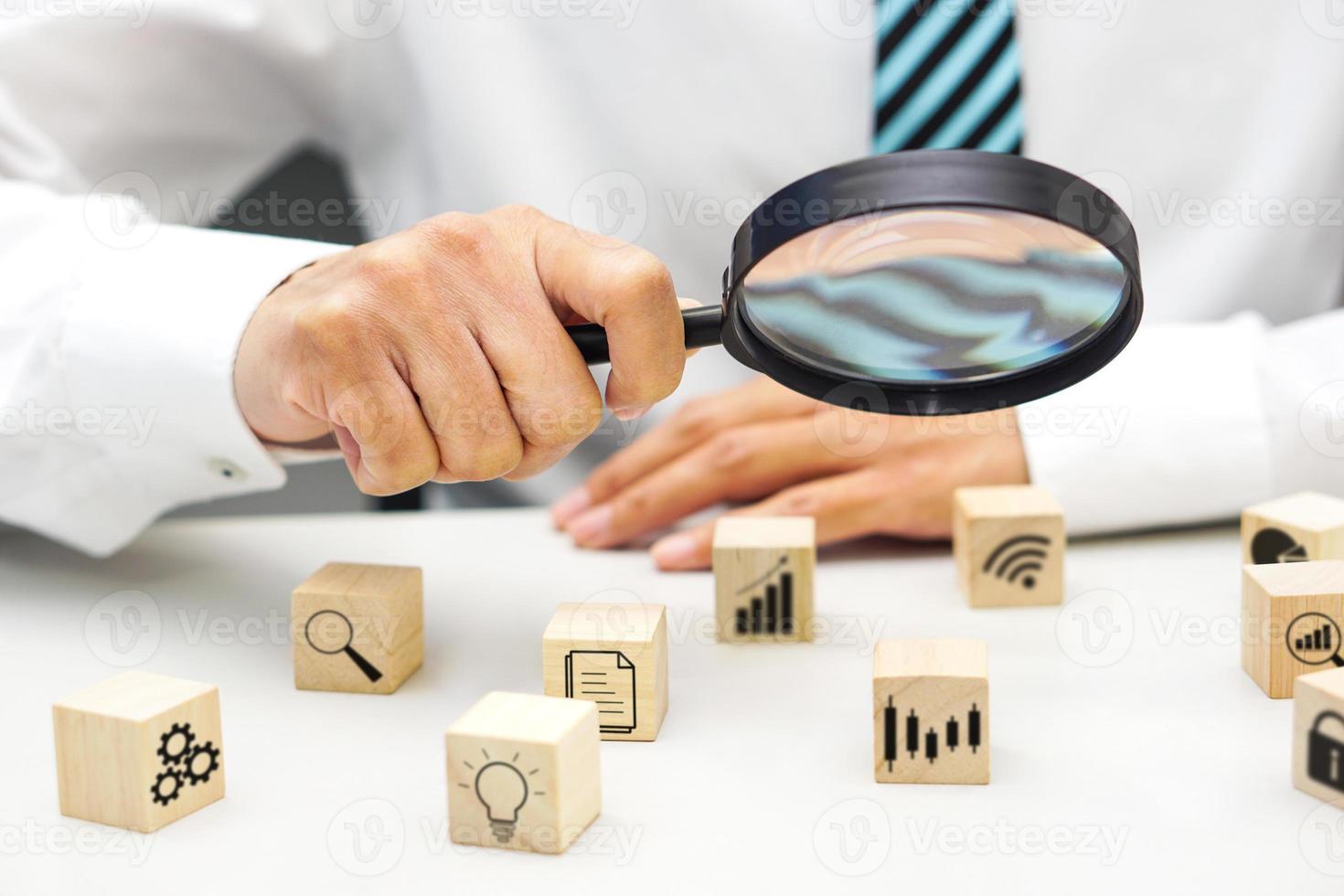 Close up hand of businessperson looking at wooden blocks with various icon symbols through magnifying glass. Concept of business strategy, goal to increase and action plan. photo