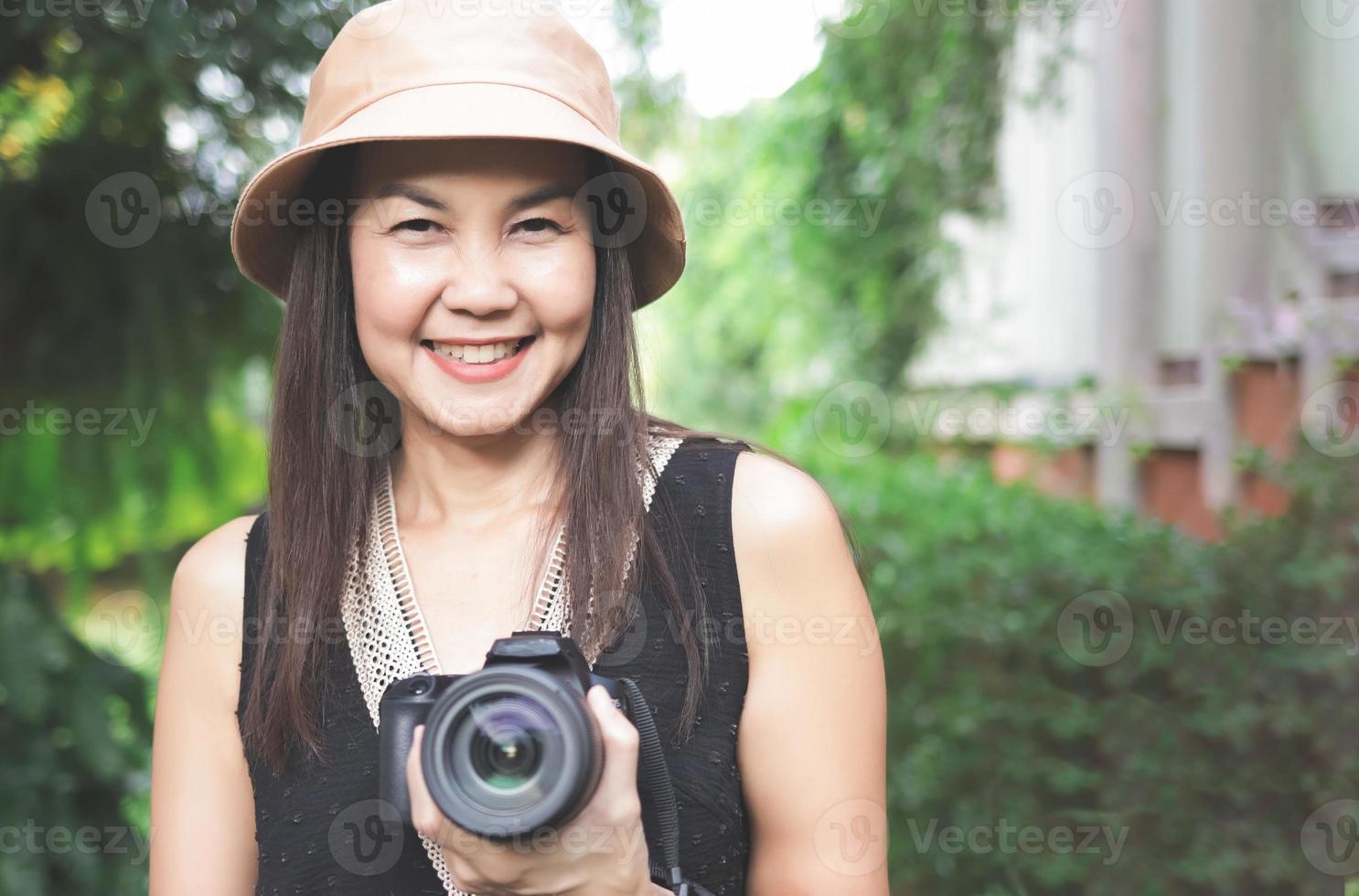 mujer asiática, con sombrero y blusa negra sin mangas, parada en el jardín y sosteniendo una cámara dslr, sonriendo alegremente. foto