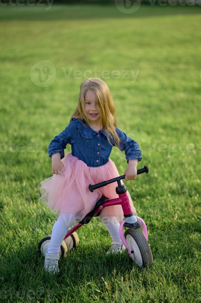 un contento hermosa pequeño niña carreras rápido y paseos su primero bicicleta sin pedales en el césped en un calentar verano día. linda bebé 3 años viejo. lado vista. aprendizaje a mantener balance. foto