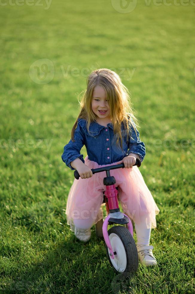 A happy beautiful little girl runs fast and rides her first bicycle without pedals on the grass on a warm summer day. Cute baby 3 years old. Side view. Learning to keep balance. photo