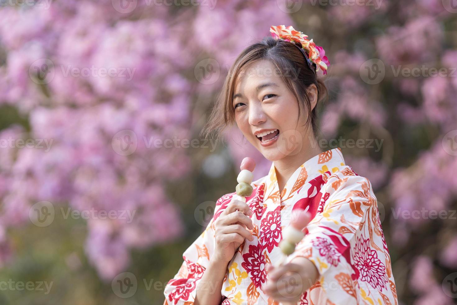 japonés mujer en tradicional kimono vestir participación dulce hanami dango postre mientras caminando en el parque a Cereza florecer árbol durante primavera sakura festival con Copiar espacio foto