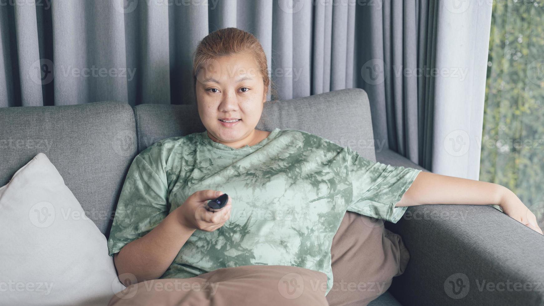asiático mujer sonrisa y contento sentar en sofá y participación remoto controlar acecho televisión con relajante acción. pasatiempo y gratis hora vida estilo concepto. foto
