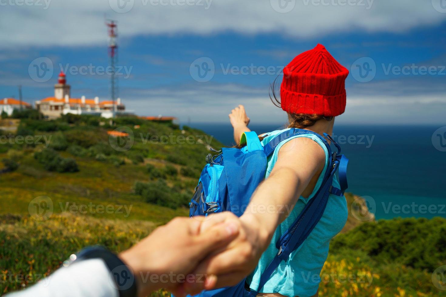 Follow me - happy young woman in a red hat and with a backpack behind her back pulling guy's hand. Hand in hand walking to the lighthouse photo