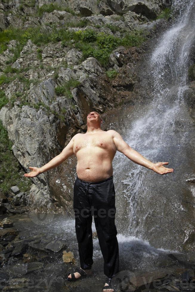 An elderly man stands at a mountain waterfall, spread his arms and enjoys the moment. photo