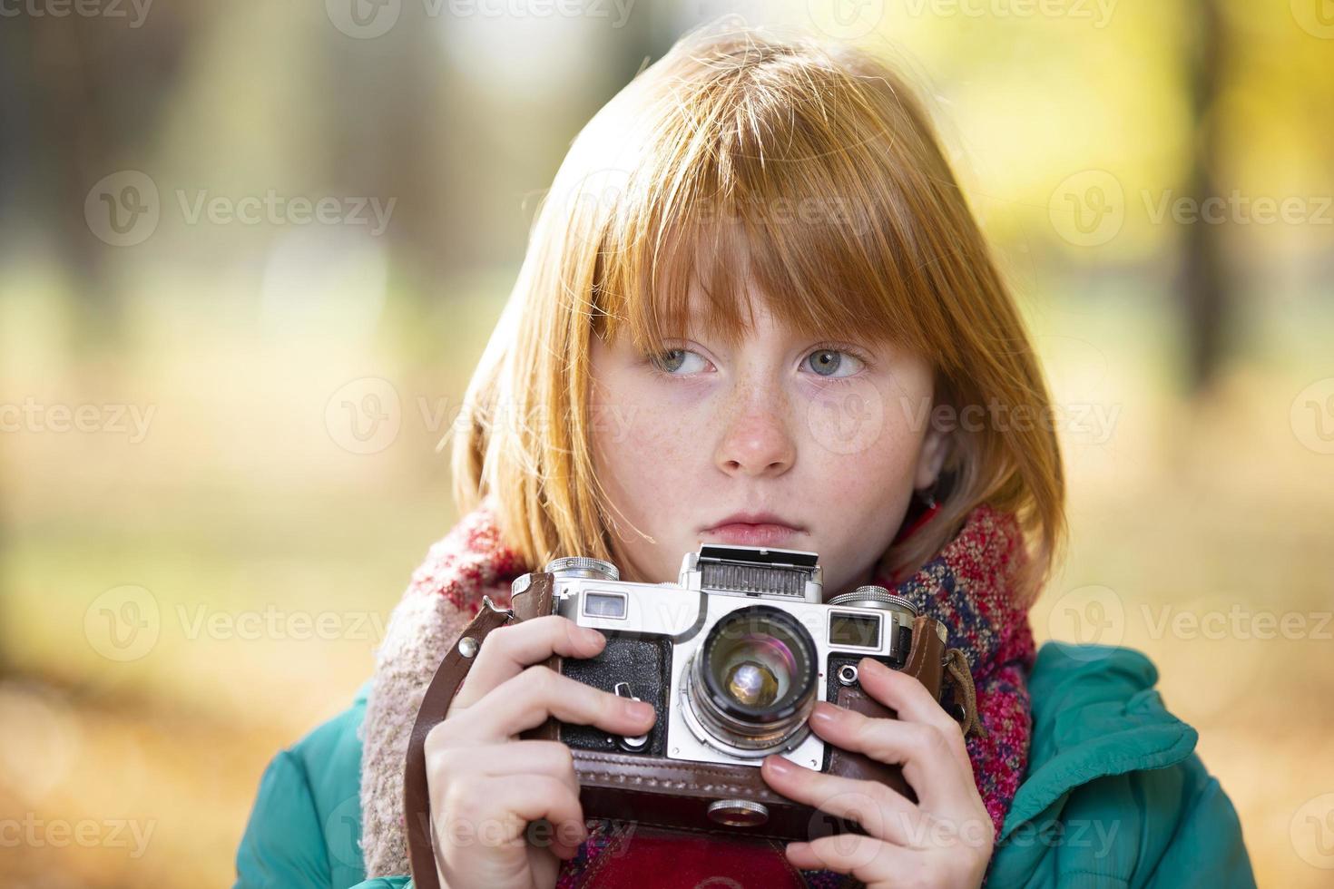 Little red-haired girl with a retro camera in the autumn park. Child photographer. photo