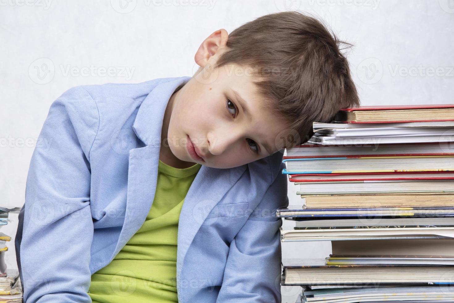 A tired school-age boy sits between stacks of books. Sad child with books. photo