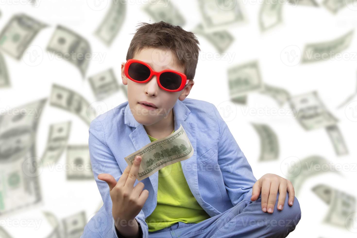 A cute, rich boy in glasses holds dollars and knows how to make money against the background of falling banknotes. photo