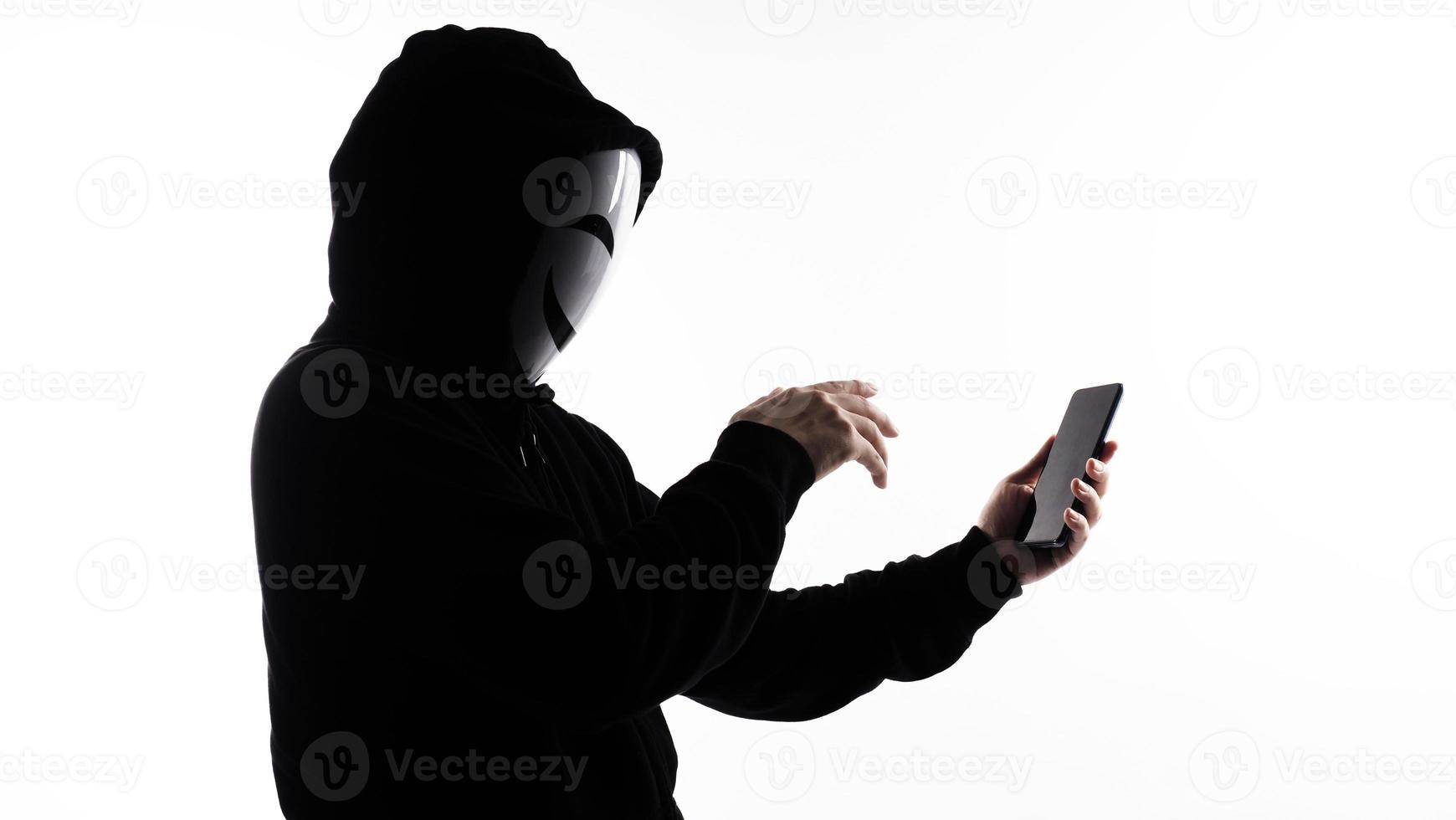 Hacker Anonymous and face mask with smartphone in hand. Man in black hood shirt holding and using mobile phone on white background. Represent cyber crime data hacking or stealing personal data concept photo