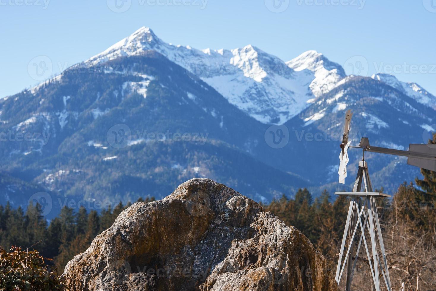 Weather vane by large rock with idyllic snow covered mountain in photo