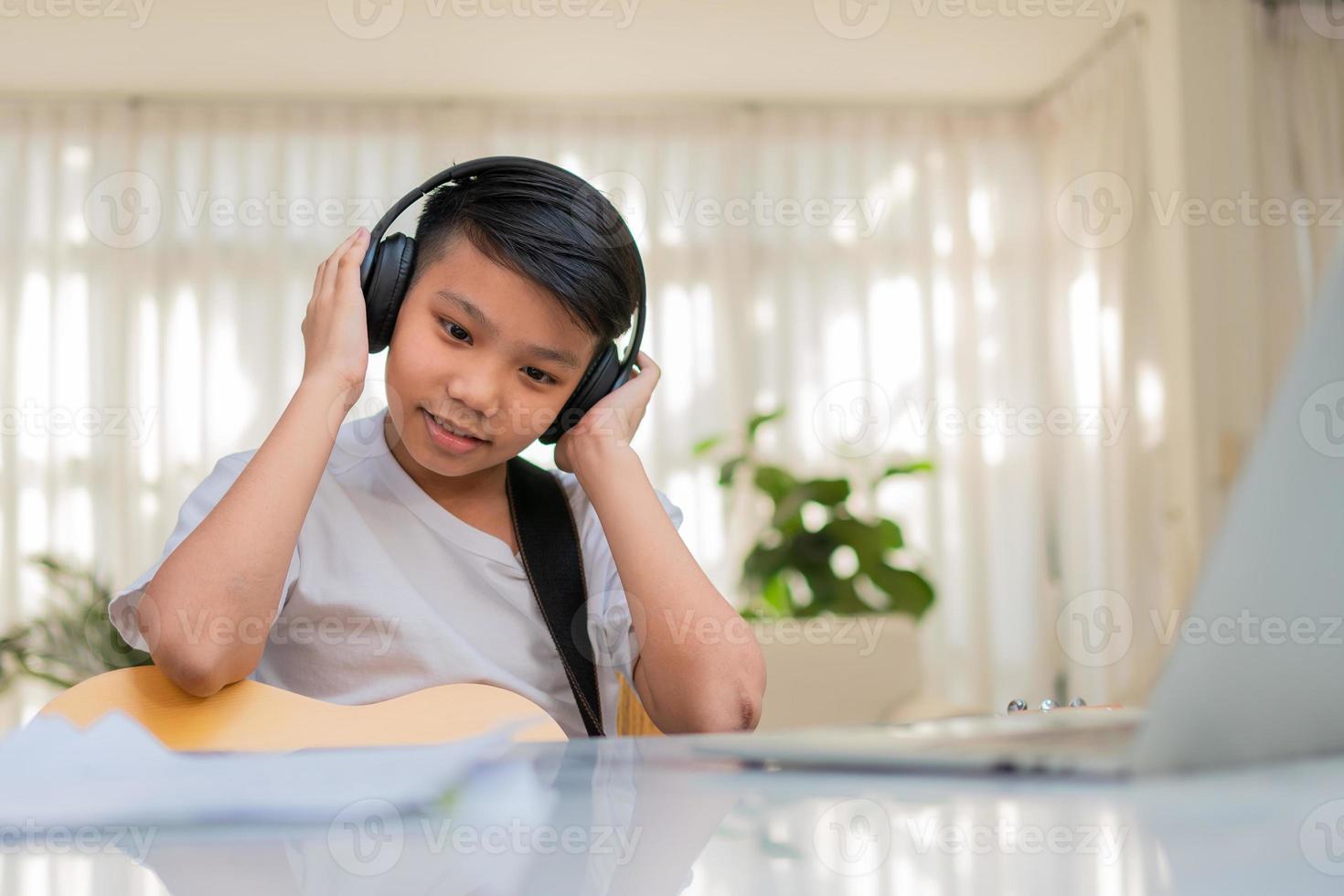 Asian boy playing guitar and watching online course on laptop while practicing for learning music or musical instrument online at home. Boy students study online with video call teachers play guitar. photo