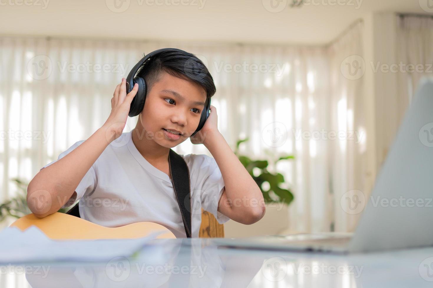Asian boy playing guitar and watching online course on laptop while practicing for learning music or musical instrument online at home. Boy students study online with video call teachers play guitar. photo