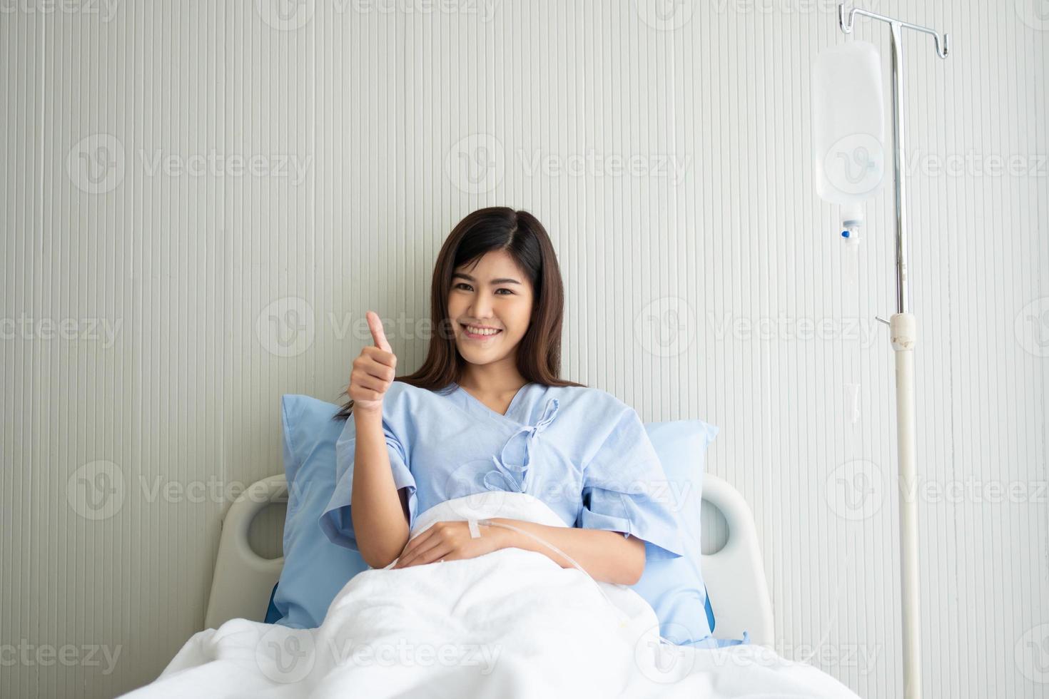 Happy Asian female patient smiling, lies on the bed, and raises thumb up to show confidence in treatment. Concept of believe in treatment And insurance coverage photo