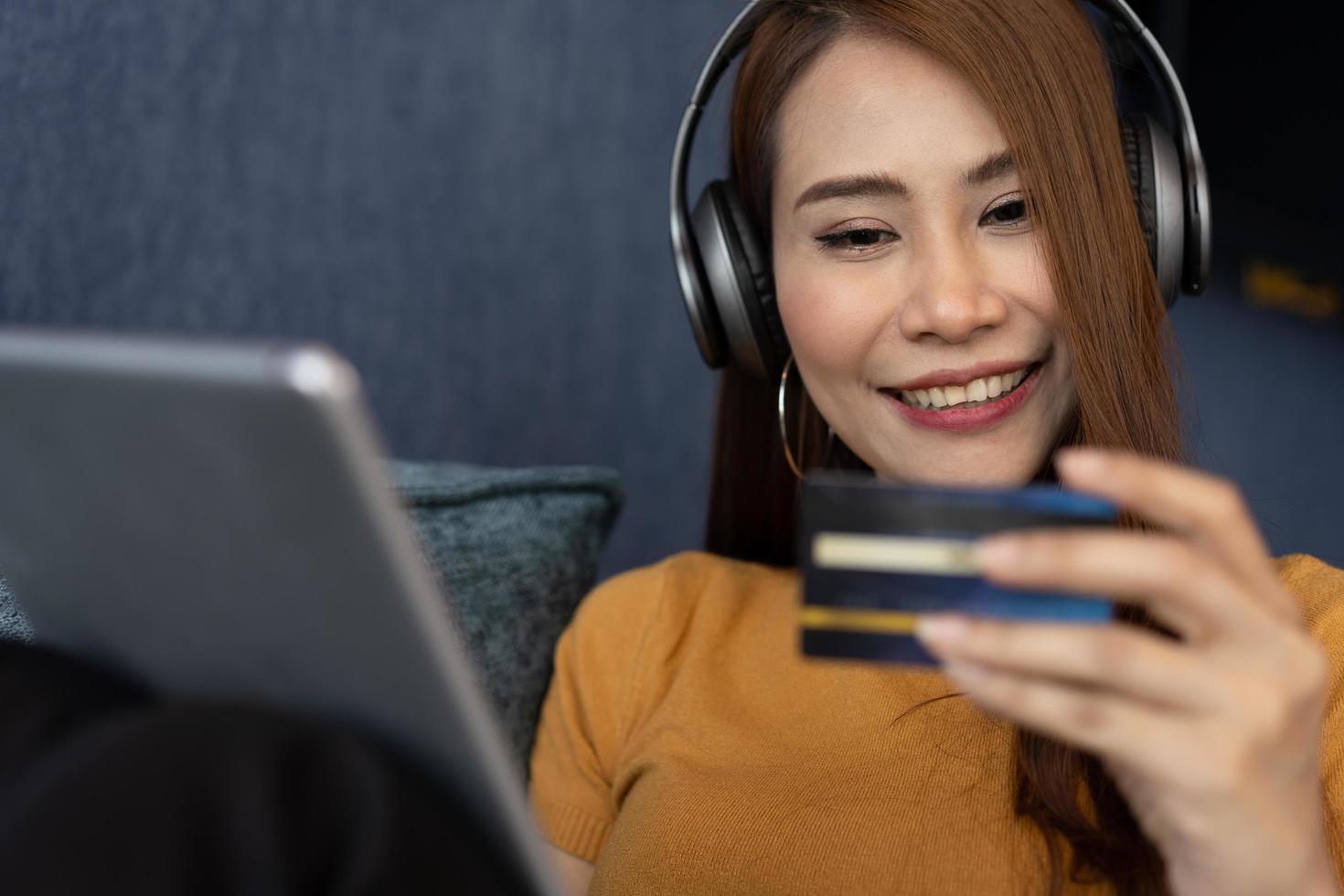 retrato de joven mujer vestir auriculares y participación tableta y bancario crédito tarjeta, transferir dinero en línea, compras bienes en Internet almacenar, adquisitivo servicios, satisfecho con seguro pagos foto