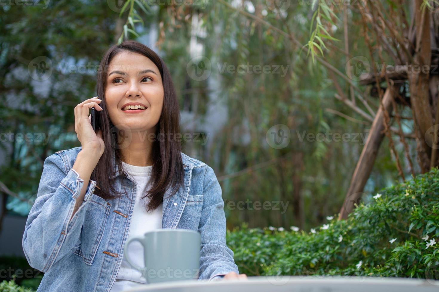 Young Asian woman freelancer holding smartphone for talking with friend. Lifestyle women working at home concept. sitting in thicket of tropical. Girl on holiday sitting in garden smiling relaxing. photo