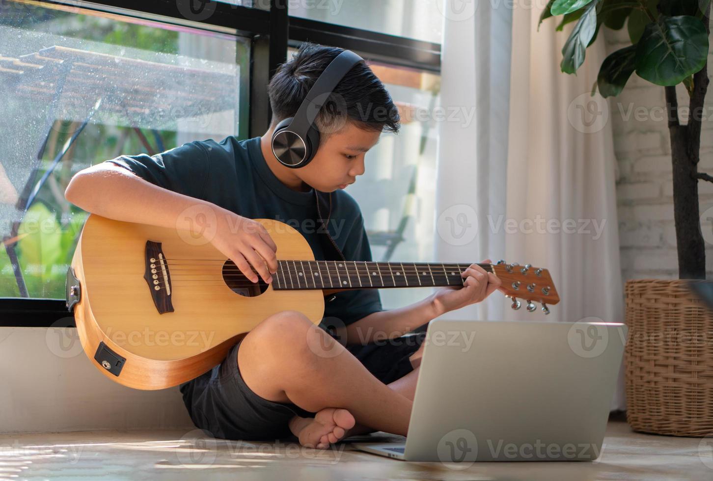 Asian boy playing guitar and watching online course on laptop while practicing for learning music or musical instrument online at home. Boy students study online with video call teachers play guitar. photo