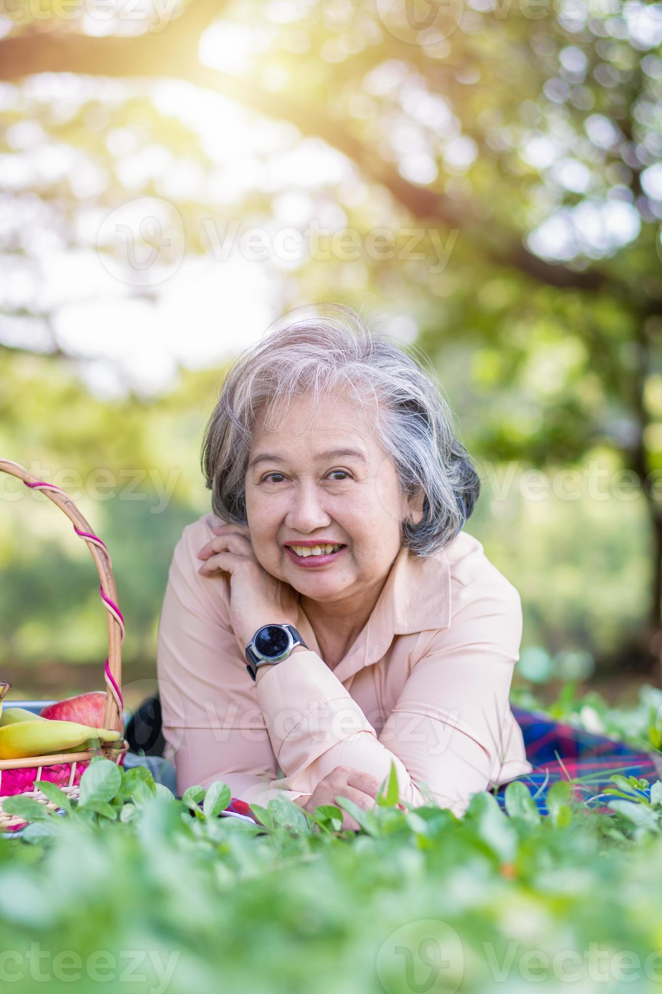 Happy Asian old senior woman and wear a health watch and lying on the  picnic mat in park and basket of fruit besides. Concept of happy elderly  woman after retirement and good