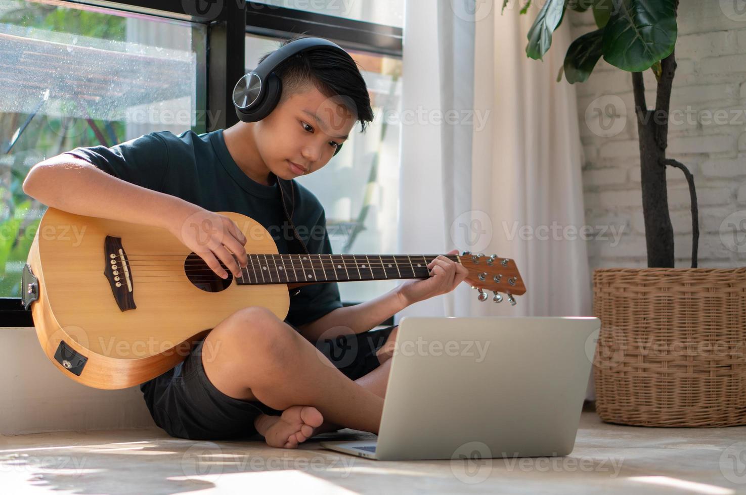 Asian boy playing guitar and watching online course on laptop while practicing for learning music or musical instrument online at home. Boy students study online with video call teachers play guitar. photo