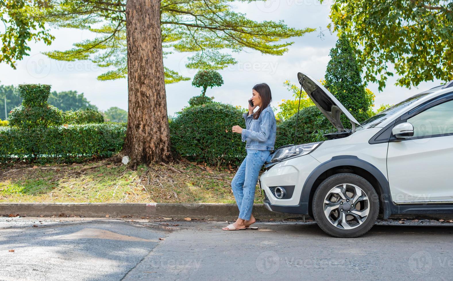 Angry Asian woman and using mobile phone calling for assistance after a car breakdown on street. Concept of vehicle engine problem or accident and emergency help from Professional mechanic photo