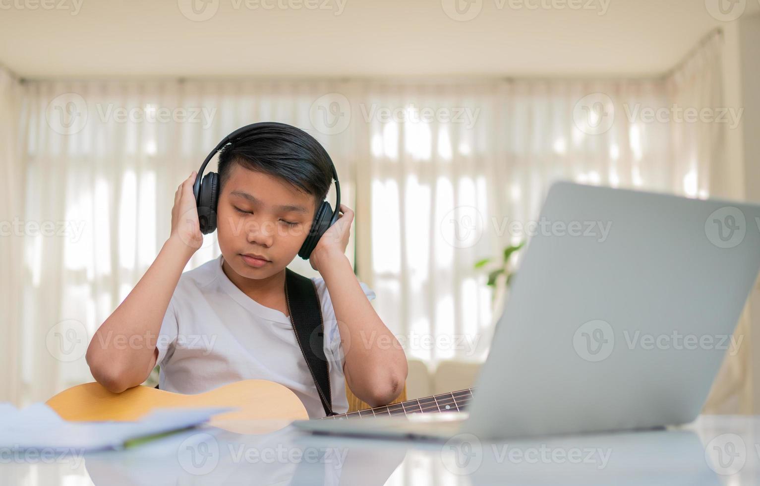Asian boy playing guitar and watching online course on laptop while practicing for learning music or musical instrument online at home. Boy students study online with video call teachers play guitar. photo