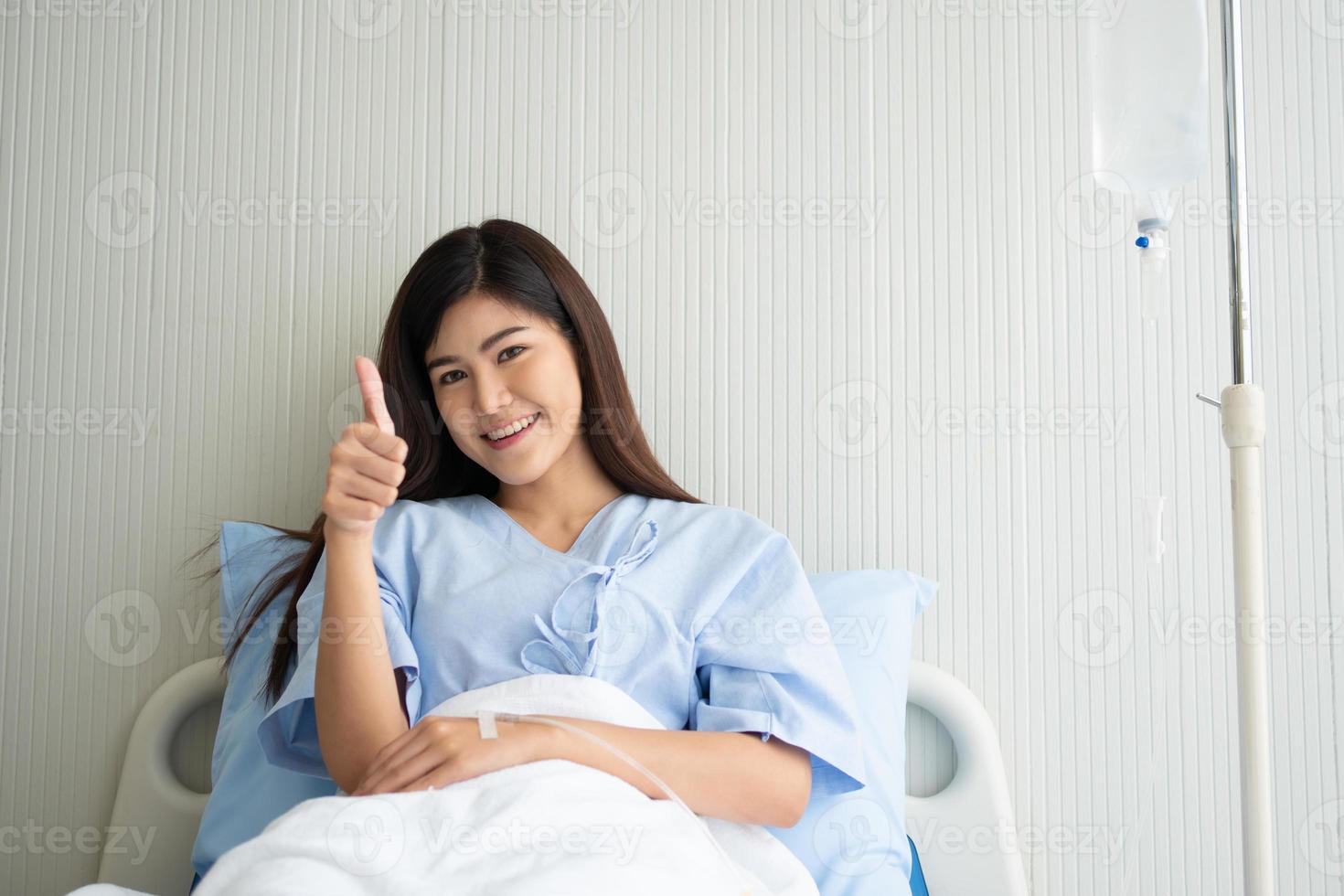 feliz paciente asiática sonriendo, se acuesta en la cama y levanta el pulgar para mostrar confianza en el tratamiento. concepto de creer en el tratamiento y la cobertura de seguro foto