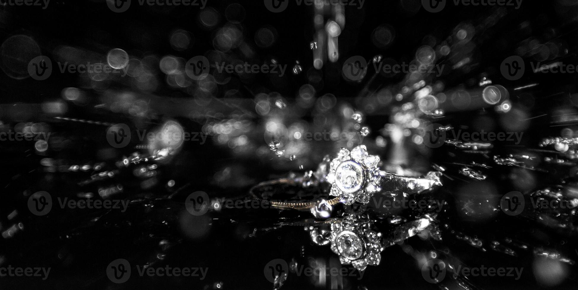 A diamond engagement and a wedding ring on a reflective surface with water splashing down from above. photo