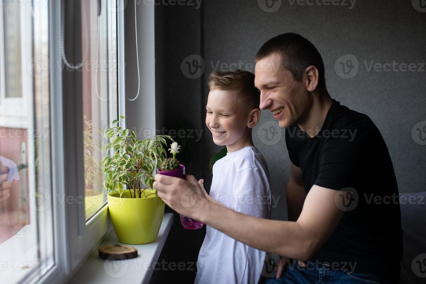A cute boy is watering indoor plants. Dad next to his son near the window photo