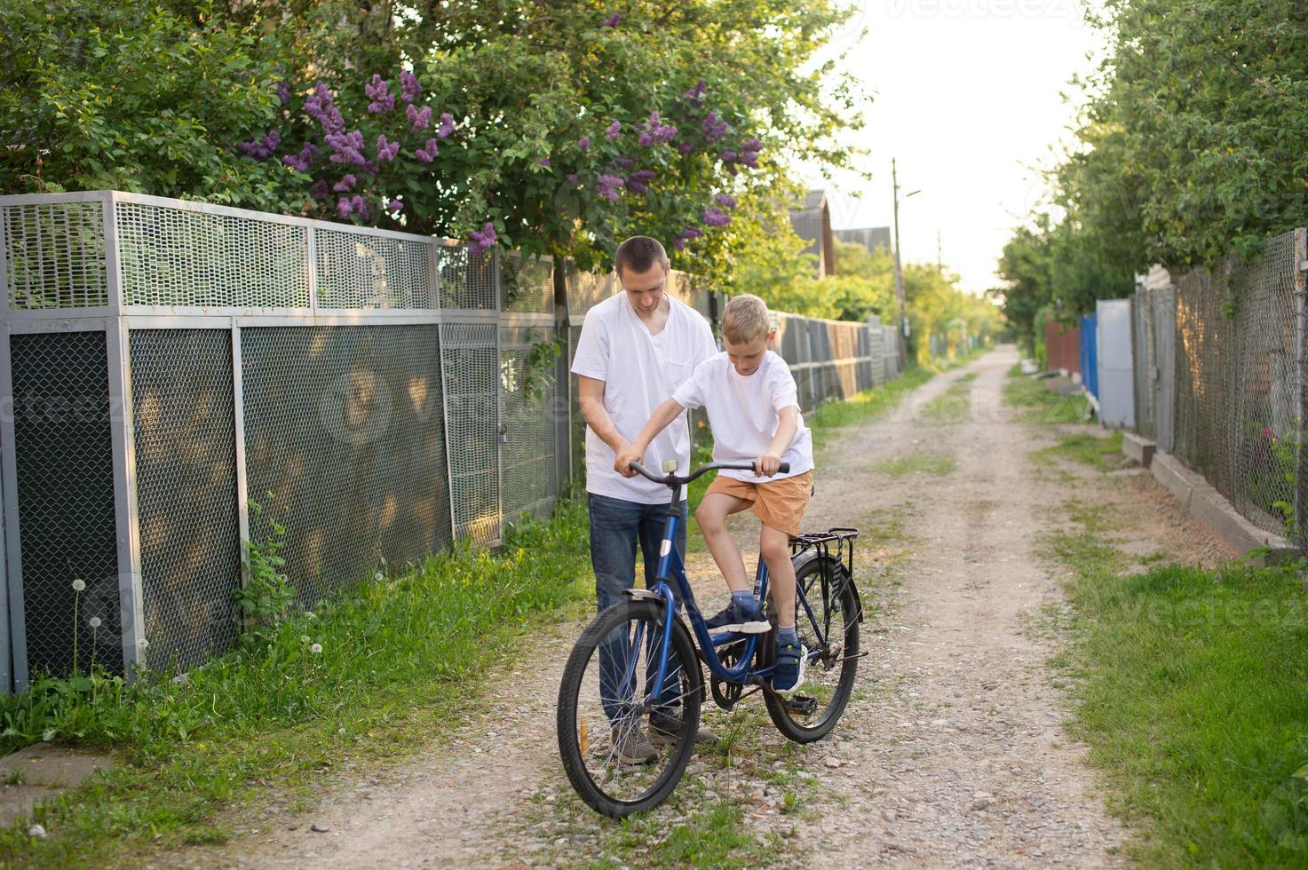A cute boy in a white T-shirt learns to ride a bike with his dad and laughs. Dad teaches his son. photo