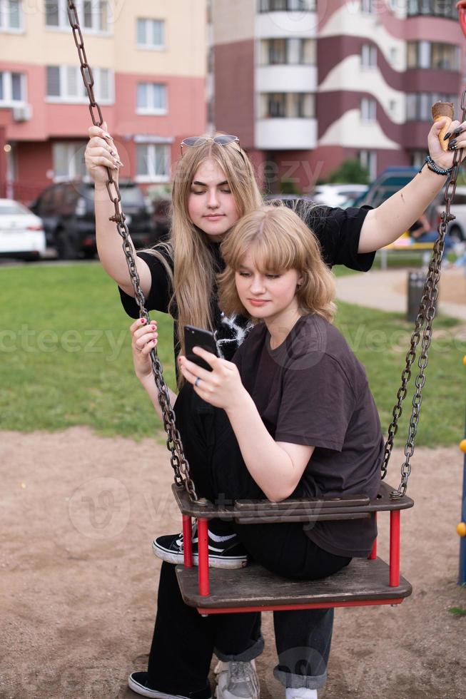 Two funny girls are sitting on a swing in the city and taking selfies on their phone. Teenagers smile photo