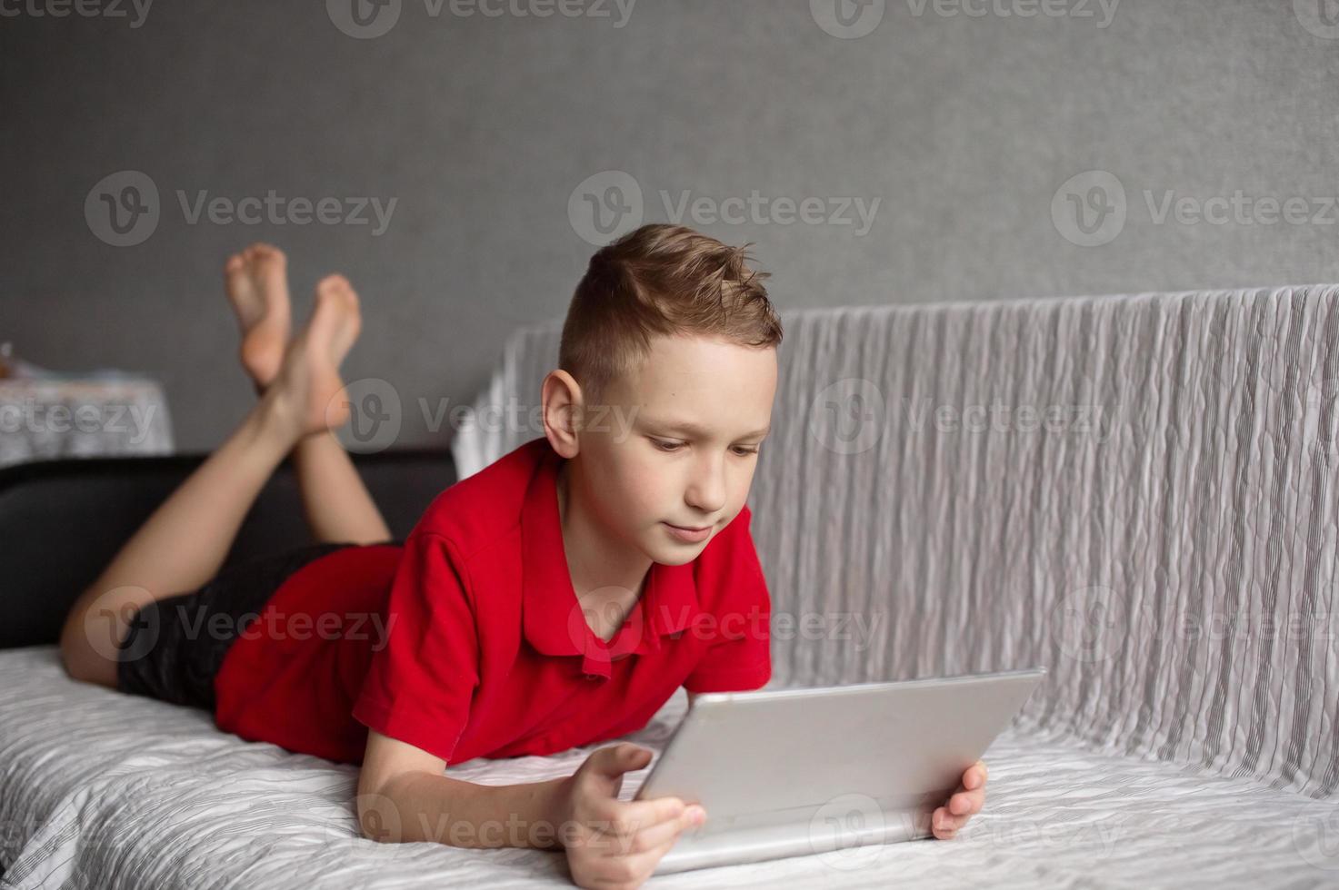 A cute boy in a red T-shirt is lying on the couch and looking at a tablet photo