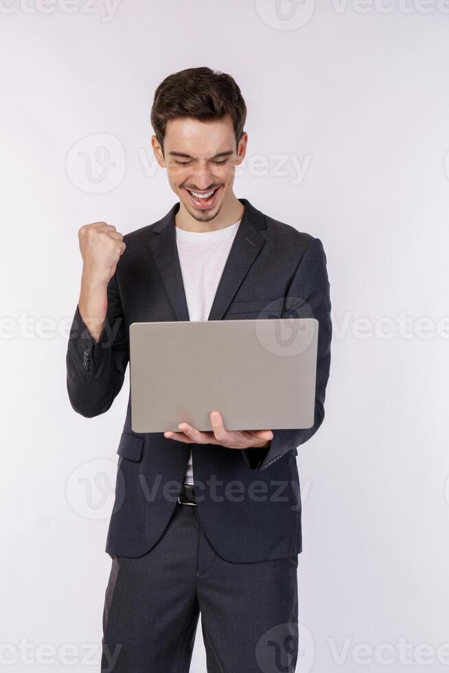 retrato de joven sonriente empresario participación ordenador portátil en manos, mecanografía y hojeada web paginas mientras haciendo un victorioso cerrado puño gesto aislado en blanco antecedentes foto