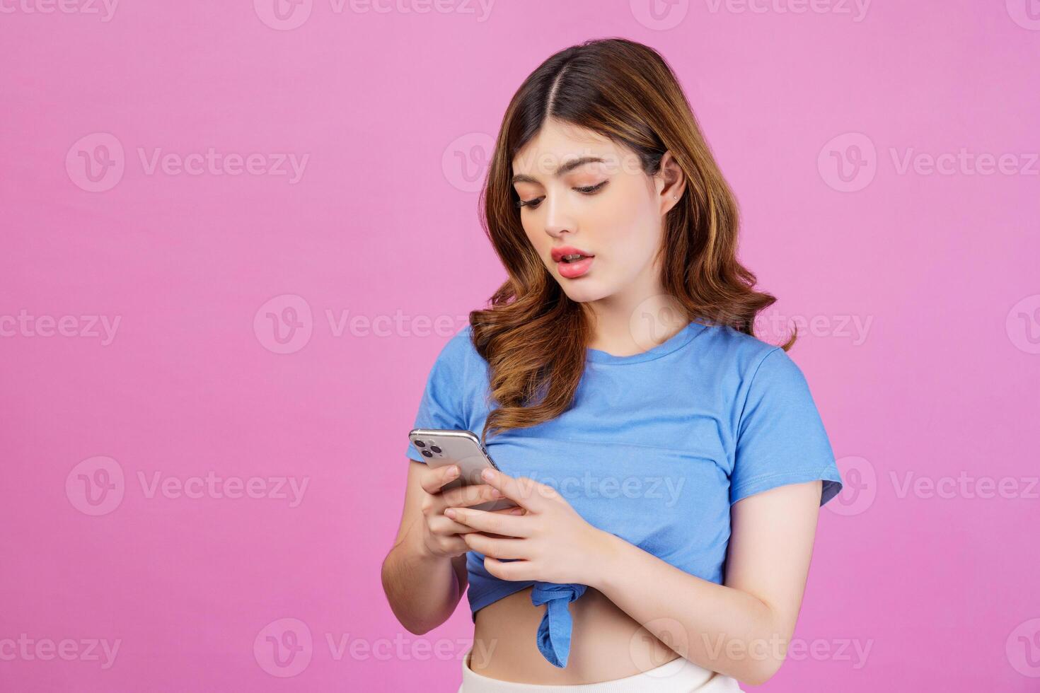 Portrait of young woman holding mobile phone in her hands while standing isolated over pink background photo