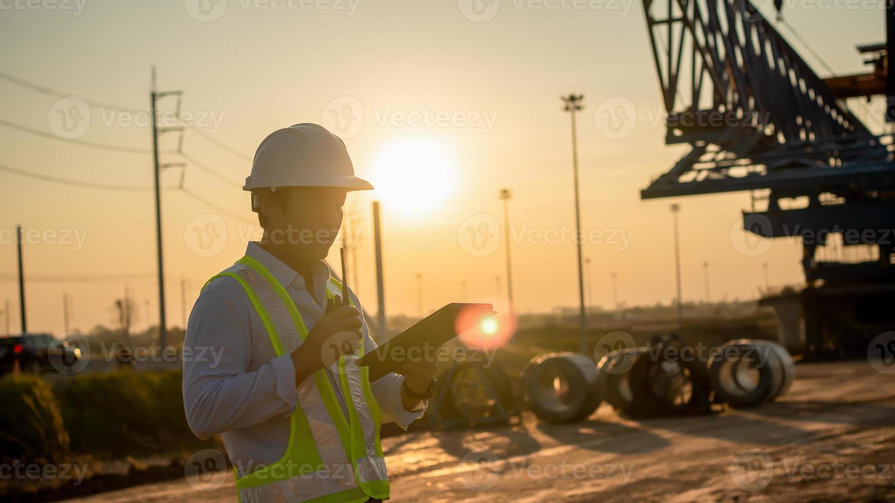 Male Engineer talking on walkie-talkie and using tablet for working at construction site on sunset photo