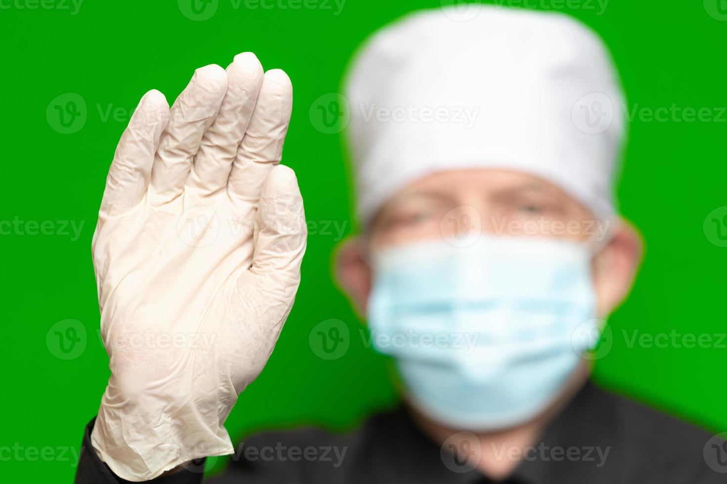 Doctor male in surgical face mask on defocus foreground put forward palm of his hand in protective latex glove foreground, waves by his hand, hello or stop sign photo