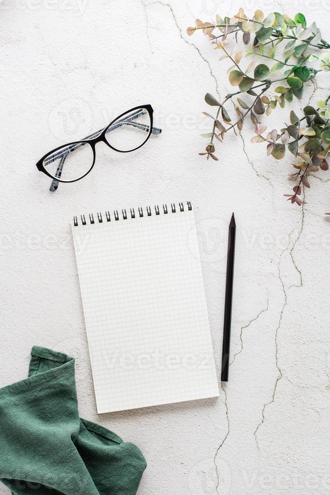 Notepad for writing, pencil, glasses and eucalyptus branch on a light textured background. Planning and organization. Top and vertical view. photo