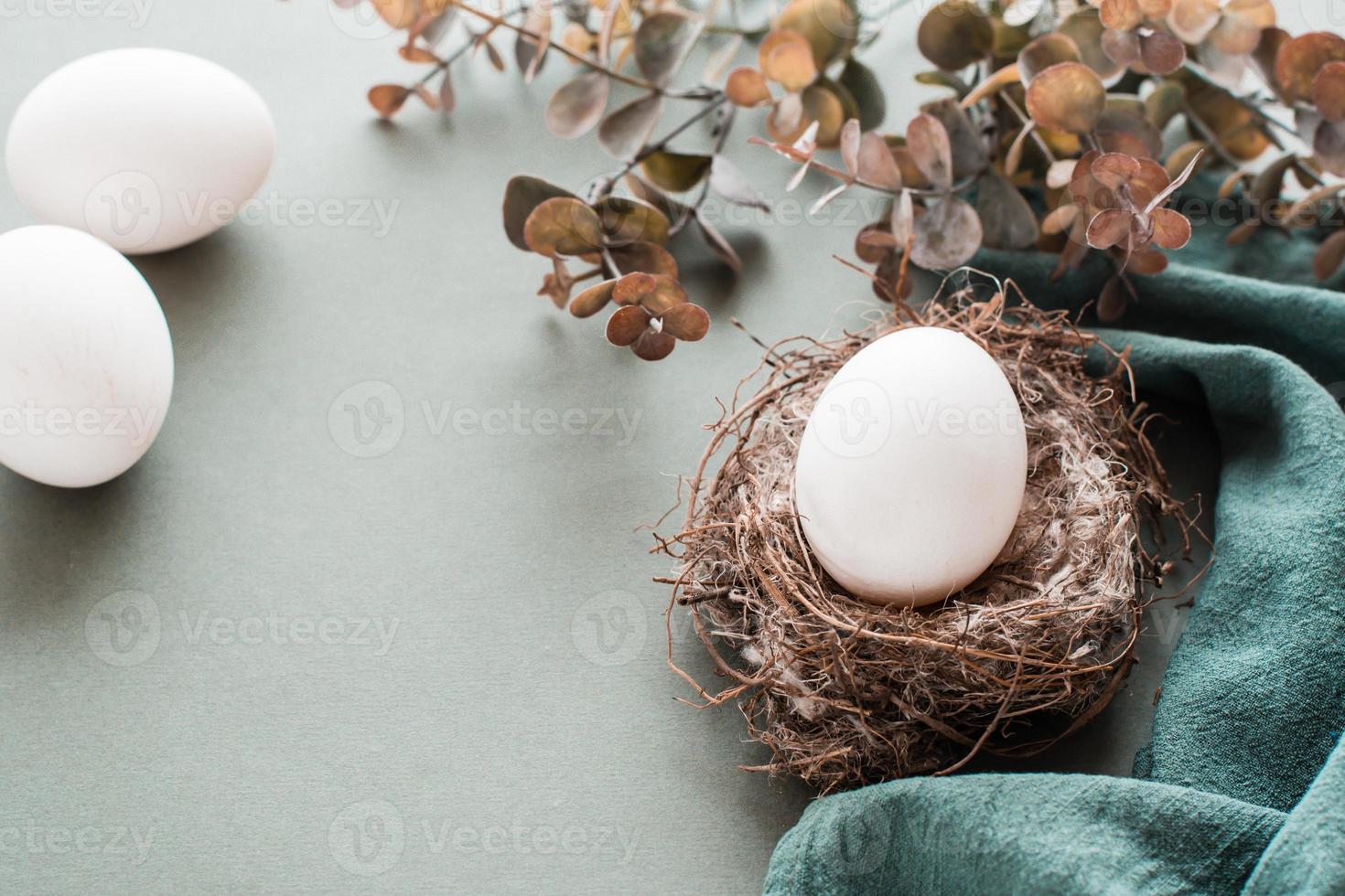 White chicken egg in a nest on a linen napkin and a branch of eucalyptus on a green background. Eco friendly happy easter. photo