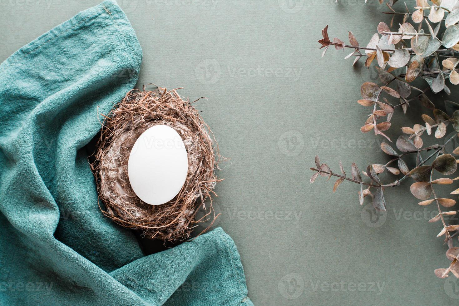 White chicken egg in a nest on a linen napkin and a branch of eucalyptus on a green background. Eco friendly happy easter. Top view. photo