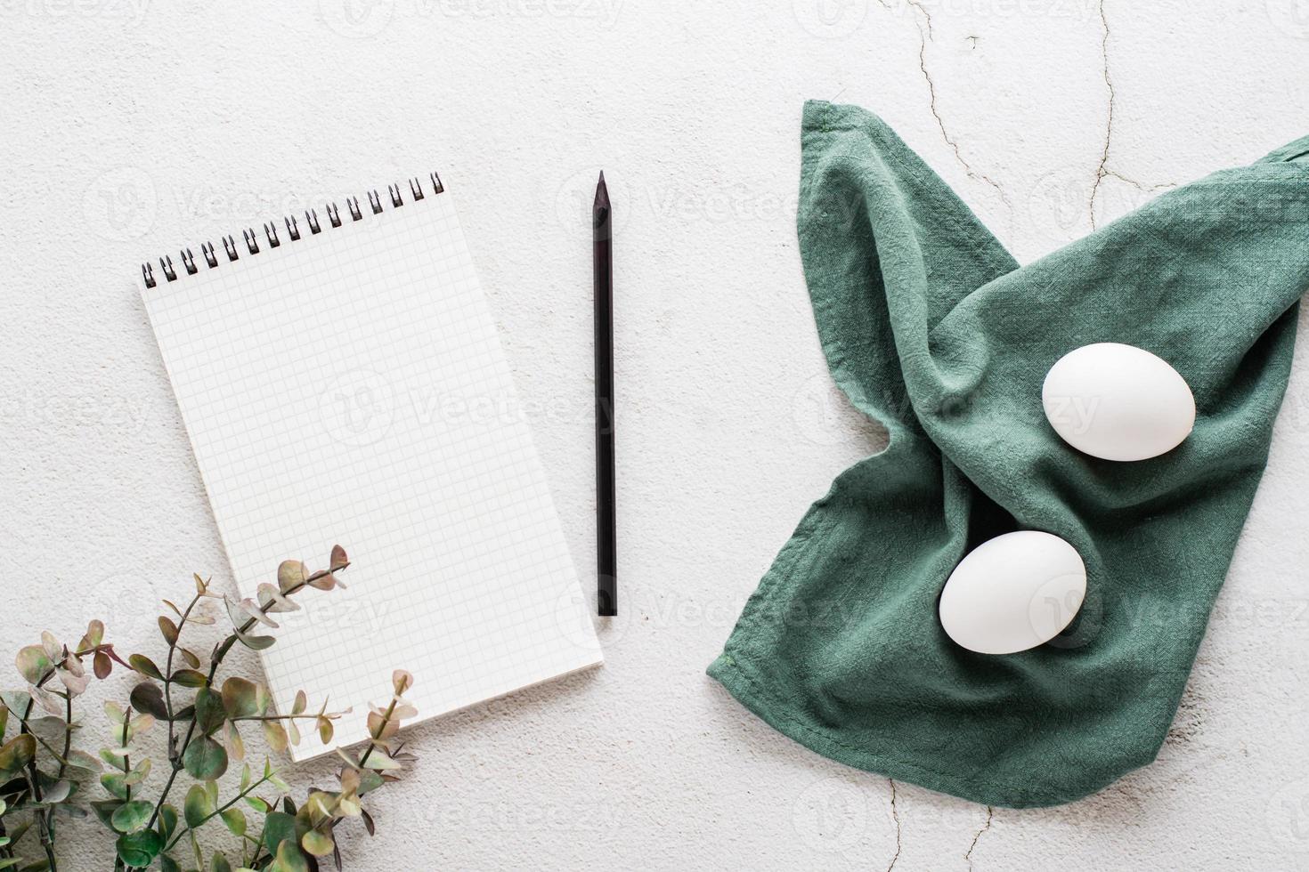 Notepad for writing, pencil, eggs on a napkin and a branch of eucalyptus on a light textured background. Happy eco easter invitation. Top view. photo