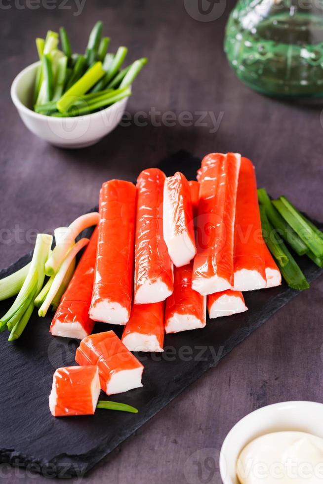 Spicy crab sticks and green onions on a stone board and sauce in a bowl on the table. Vertical view photo