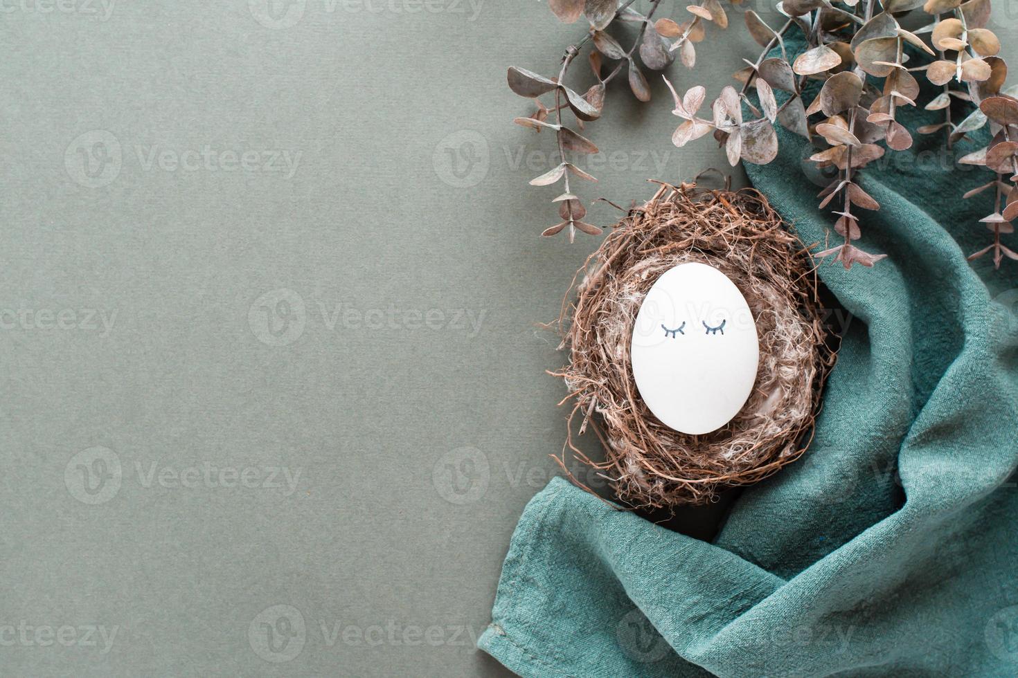 A white chicken egg with eyes in a nest on a linen napkin and a branch of eucalyptus on a green background. Eco friendly happy easter. Top view. Copy space photo