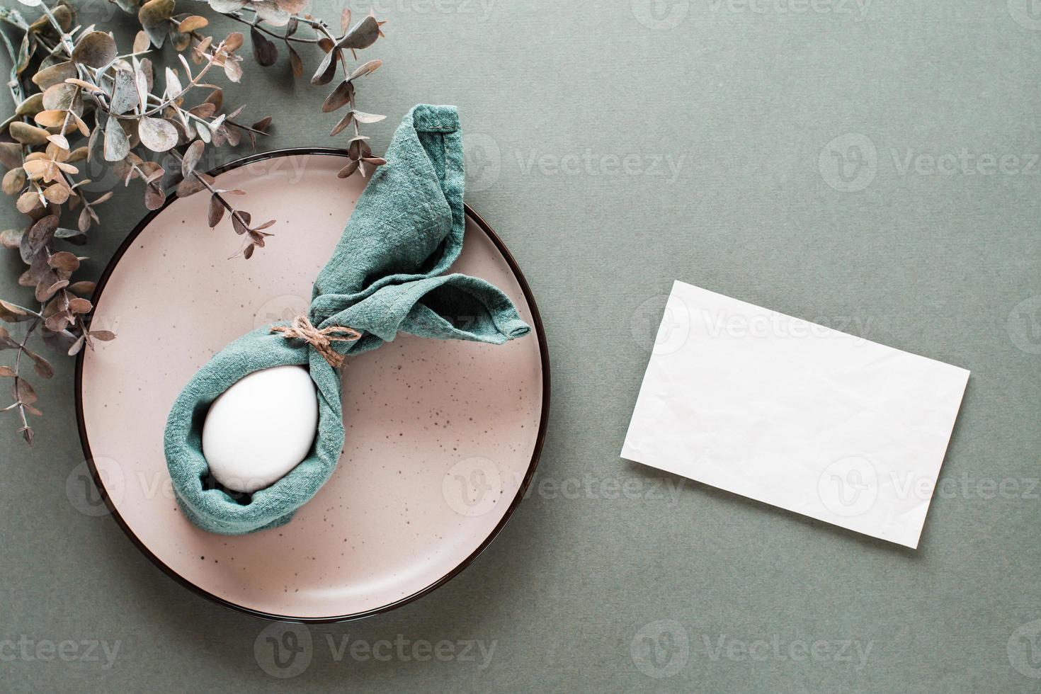 A white chicken egg is wrapped in a napkin in the form of a rabbit on a plate, a piece of paper for writing and a branch of eucalyptus on a green background. Eco friendly happy easter. Top view. photo