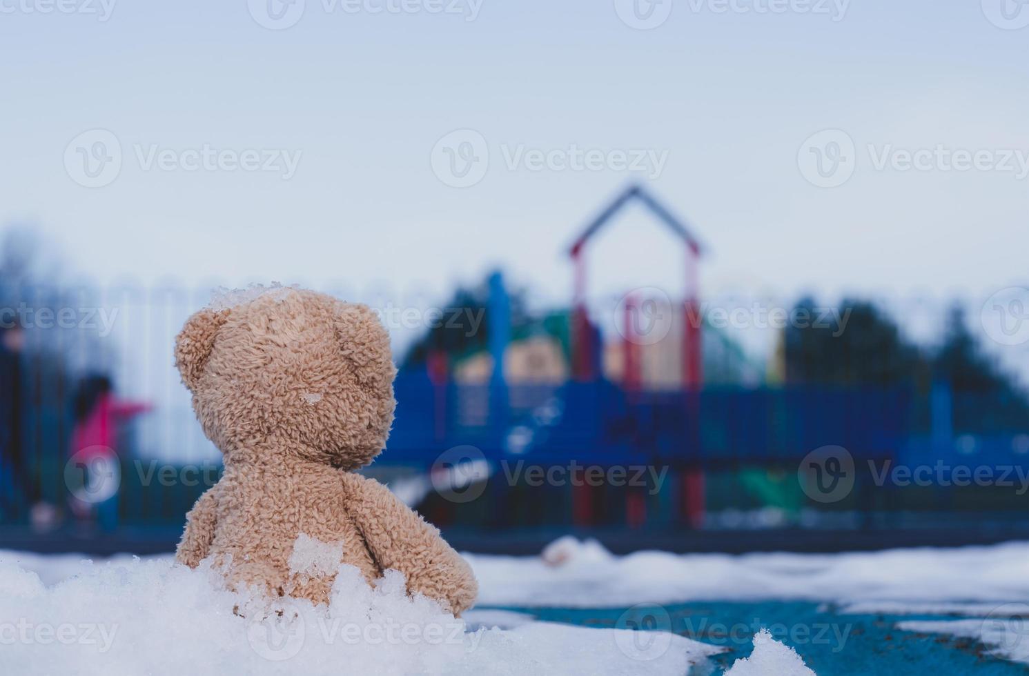 Lonely Teddy bear doll sitting alone on footpath with blurry kid playing playground in retro filter, Back view Rear view Lost brown bear toy looking at people, International missing children's day photo